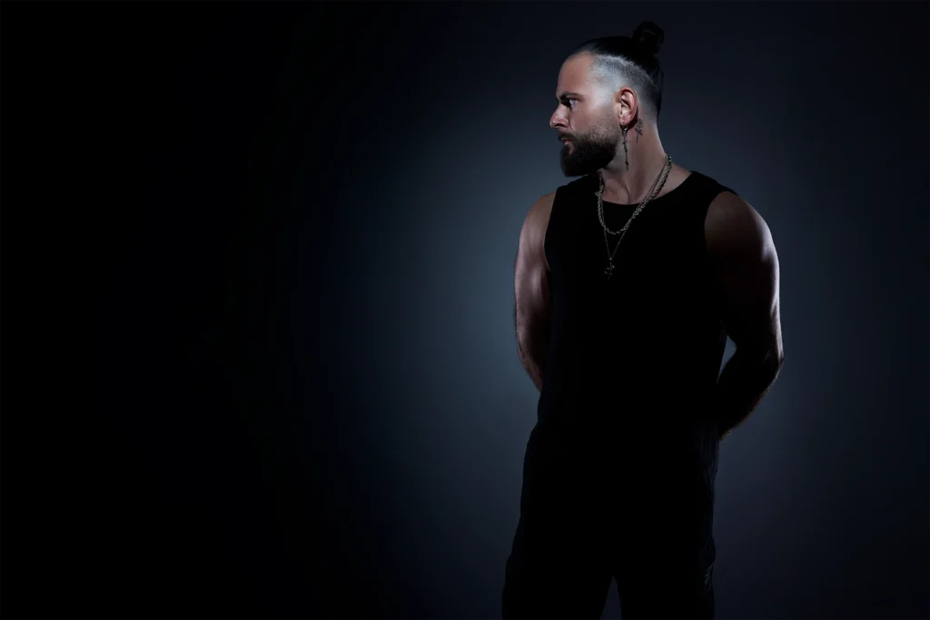 DJ Lahox de perfil con moño y camiseta negra en estudio, foto de marca personal.